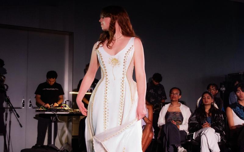 Model walks in a white dress featuring gold embroidery and a sunburst motif on the bodice. The structured gown has a sweeping hemline and is framed by seated spectators.